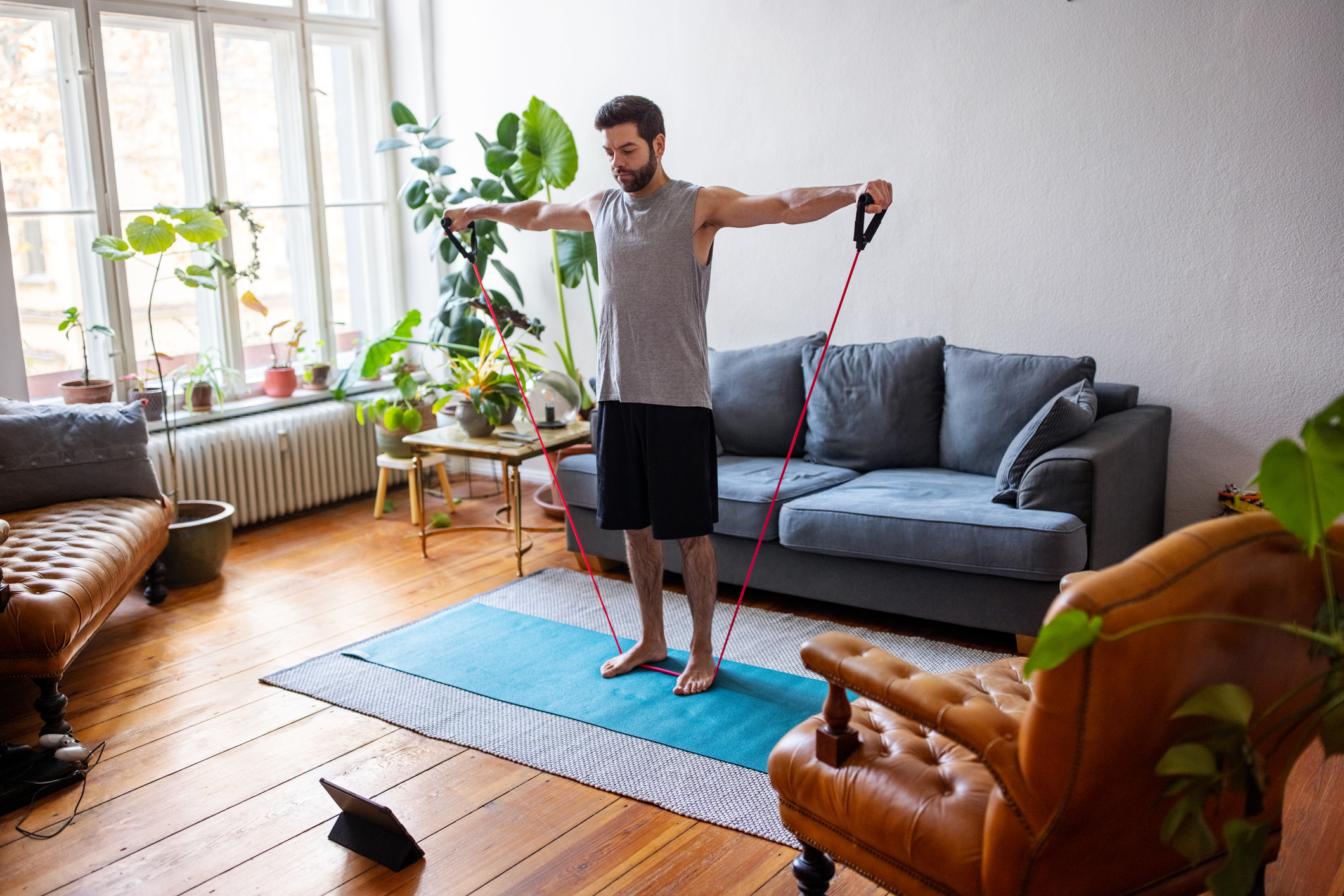 Man exercising with stretching band at home with a digital tablet. Mid adult man using an elastic rubber band following a tutorial online and exercise at home during isolation for Covid - 19 at home.