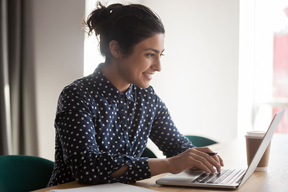 Smiling indian female employee busy working on laptop typing writing business letter to client, positive ethnic woman worker message on computer browsing internet or checking mail online in office