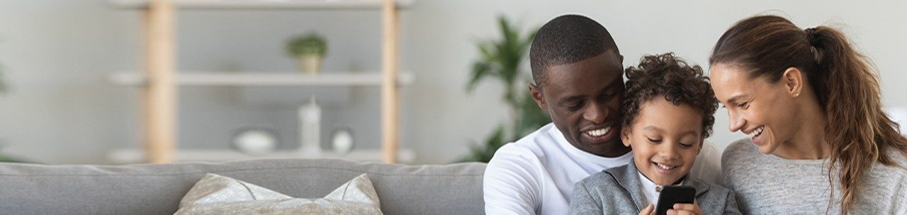 Smiling young woman sitting at living room on comfortable couch near happy african american husband holding cute small mixed race boy on knees, watching little kid son playing on mobile phone.