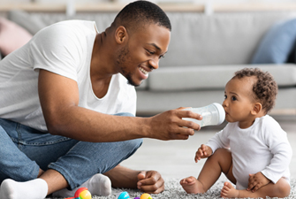 Caring young black dad giving bottle with water to his little baby at home, loving african american father babysitting cute adorable toddler child, playing together on floor in living room