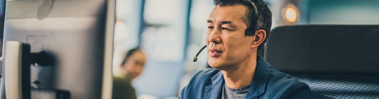 Photo of a mature Asian man with a headset using a computer while working in a call centre.