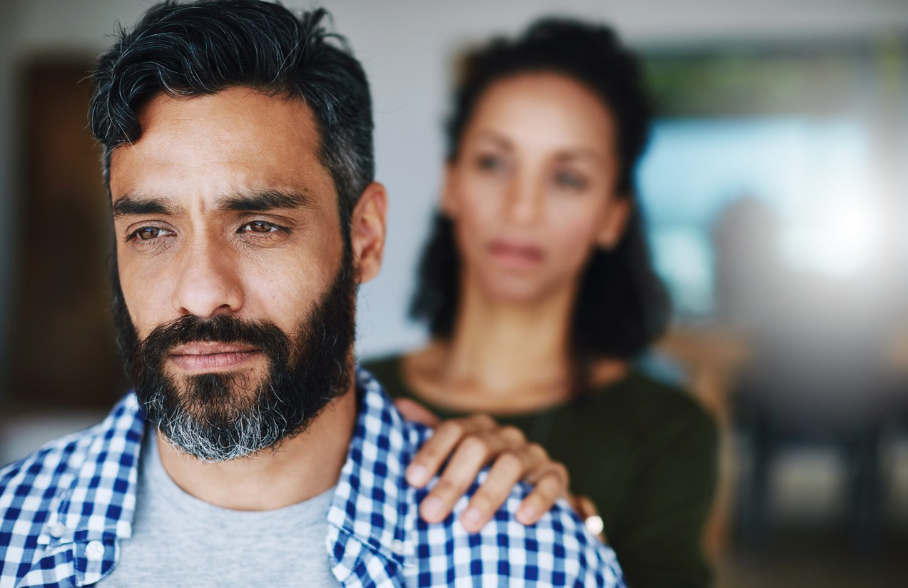 A somber-looking man stares off while a woman (blurred in the background) places a comforting hand on his shoulder.